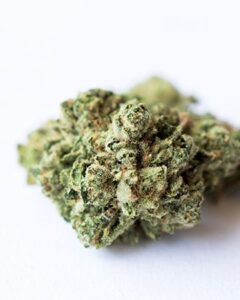 A Runtz Seeds cannabis bud sits idly on a plain white surface, showcasing dense, green clusters with hints of orange and a frosty coating of trichomes.