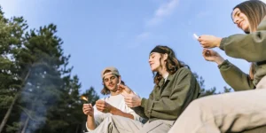 Three friends relaxing by a campfire under a blue sky, holding small lights.