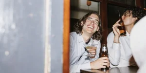 Group of friends at a lively bar table, one laughing with a martini, another drinking from a beer bottle.